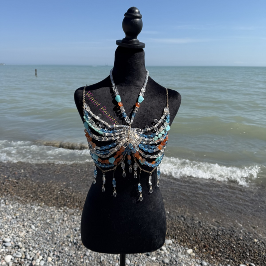 Translucent blue, orange, and clear crystal beaded body chain bra with gold fringe accents on a black mannequin at the beach.