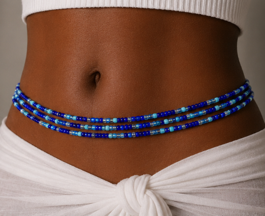A close-up of a person's waist wearing a beaded waist chain made of blue and light blue beads, styled with a white ribbed crop top and a white knotted fabric at the waist.