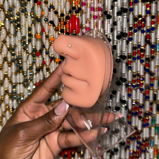  A close-up photograph of a realistic tan silicone nose display model with a single round clear crystal stud piercing, held in a hand with French manicured nails against a colorful beaded curtain background.