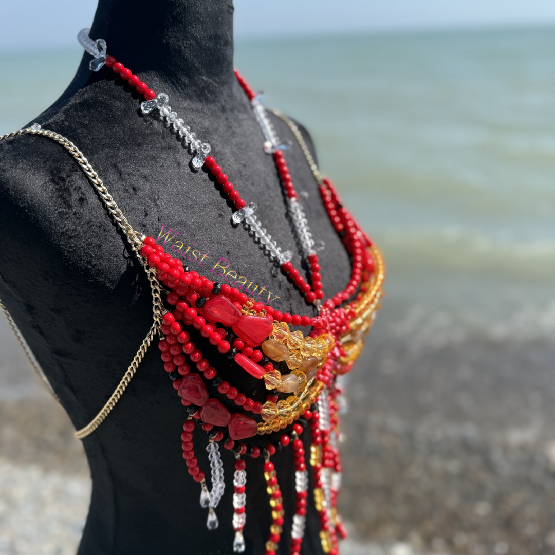 Waist Beauty - Beaded Bra - Vibrant red and amber gold crystal beaded festival bra with long underbust fringe on a black mannequin with a beach background.