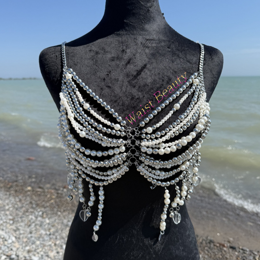 Metallic silver and pearl layered body chain bra with dangling heart charms displayed on a black mannequin at a rocky beach.
