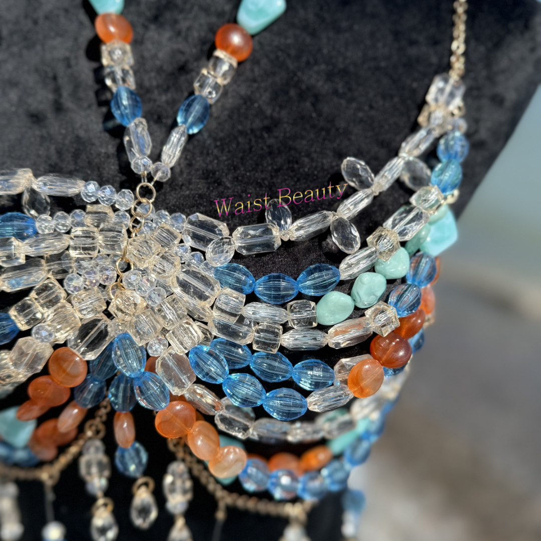 Translucent blue, orange, and clear crystal beaded body chain bra with gold fringe accents on a black mannequin at the beach.