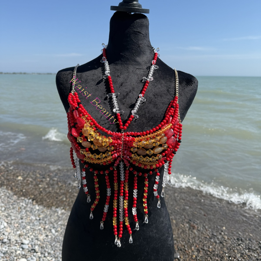 Vibrant red and amber gold crystal beaded festival bra with long underbust fringe on a black mannequin with a beach background.