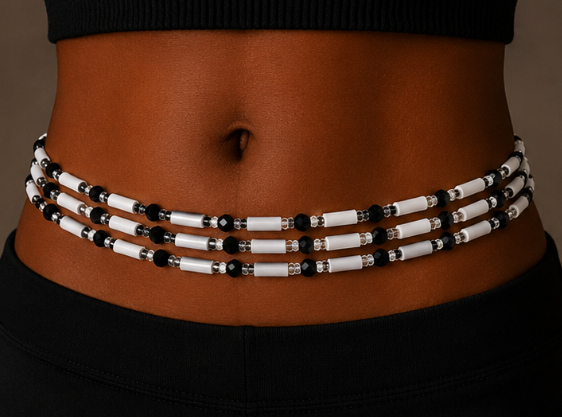 A close-up of a model's waist wearing a multi-strand beaded waist wrap. The wrap features alternating white cylindrical beads, black round beads, and small silver metallic spacer rings, set against a dark black top and pants.