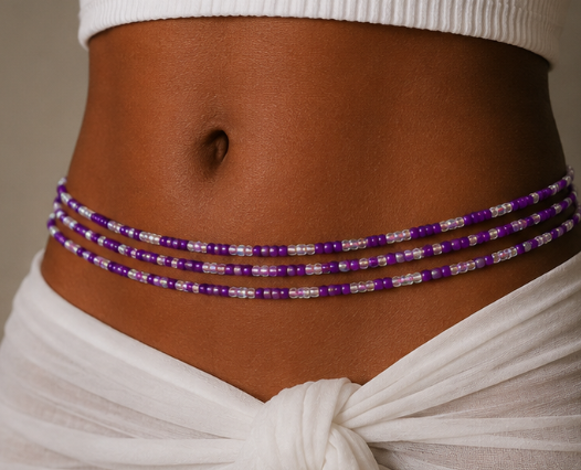 A close-up of a model's midsection wearing a multi-strand beaded waist chain with purple and clear faceted beads, styled with a white ribbed crop top and a white knotted fabric wrap.