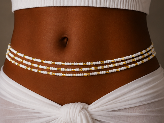 A close-up of a model's midsection wearing a beaded waist chain with white and gold beads, styled with a white wrap skirt.